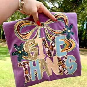 May include: A purple t-shirt with the words "GIVE THANKS" in a decorative font. The letters are in various colors and patterns, including floral and gold accents. A large bow and flowers decorate the top of the shirt.