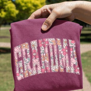 May include: A maroon-colored sweatshirt with the word "Grandma" in a floral design. The letters are filled with pink, white, and orange flowers. The sweatshirt is held up by a hand, with a blurred green background.