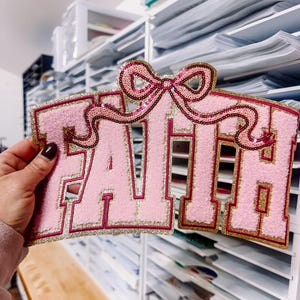 Puede incluir: Parche de tela rosa con la palabra "FAITH" en letras grandes. Las letras están delineadas en dorado y rosa oscuro, con un lazo de lentejuelas. El parche tiene una textura difusa.