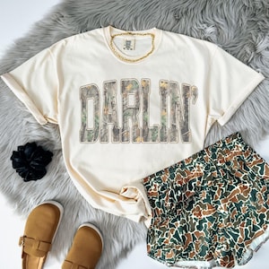 Könnte beinhalten: Cremefarbenes T-Shirt mit dem Wort "DARLIN'" in einem Camouflage-Muster. Das Shirt wird mit passenden Camouflage-Shorts, braunen Slipper-Schuhen und einer goldenen Halskette kombiniert. Ein schwarzes Scrunchie ist ebenfalls sichtbar.