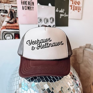 Może przedstawiać: Czapka typu trucker z białym panelem przednim, brązowym daszkiem i szarą siatką z tyłu. Czapka zawiera napis "Yeehaws & Hellnaws" czarną czcionką. Czapka jest prezentowana na lustrzanej kuli dyskotekowej.