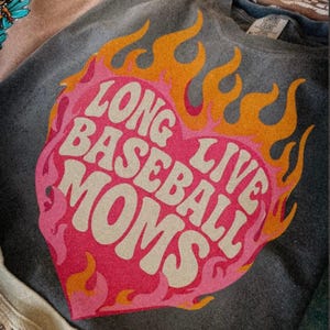 May include: A gray t-shirt with a pink heart-shaped graphic and orange flames. The text "LONG LIVE BASEBALL MOMS" is printed in white within the heart. The shirt is folded, with other shirts visible in the background.