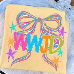 Op de afbeelding: Lichtgeel T-shirt met een groot, kleurrijk strikdesign en de tekst "WJD" in heldere letters. Het ontwerp bevat sterren in verschillende kleuren, wat een speelse en opvallende grafiek creëert.