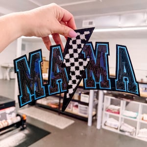May include: A large embroidered patch featuring the word MAMA in block letters with a black and blue sequined finish. A central lightning bolt shape with a black and white checkered pattern separates the letters. The patch has a black border.