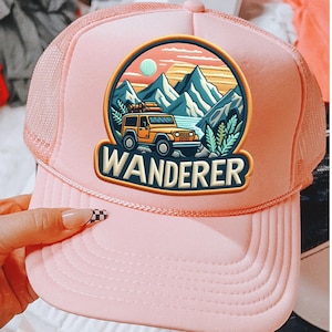 May include: Pink trucker hat with a mesh back and a curved brim. The front features a circular patch with a yellow jeep, mountains, and the word "WANDERER". The patch has a blue, orange, and green color scheme.