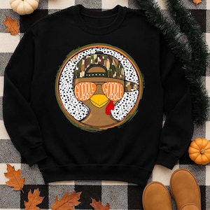 May include: Black sweatshirt featuring a turkey wearing a camouflage hat and sunglasses. The sunglasses have the words "GOBBLE GOBBLE" printed on them. The turkey is inside a white polka dot circle with a green and orange border. A Thanksgiving theme.