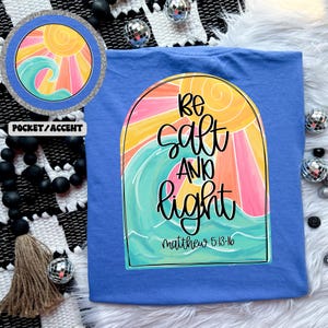 May include: Blue t-shirt with a colorful graphic featuring a sun, waves, and the words "Be Salt and Light" in black script. The text "Matthew 5:13-16" is below the main text. A smaller version of the graphic is in the upper left corner.