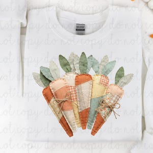 Puede incluir: Sudadera blanca con un diseño de zanahorias coloridas. Las zanahorias están hechas de telas con patrones en naranja, verde y beige. El diseño está centrado en la parte delantera de la camisa, con cuello redondo y la marca "Gildan".