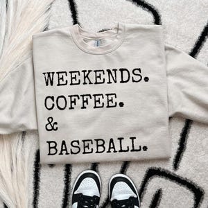 May include: A beige sweatshirt with the words "WEEKENDS. COFFEE. & BASEBALL." printed in black. The sweatshirt is paired with black and white sneakers, resting on a patterned rug. The overall theme is casual wear.