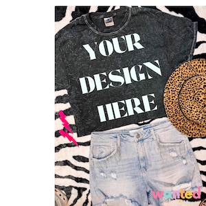 May include: A black cropped t-shirt with the text "YOUR DESIGN HERE" in white. The shirt is on a zebra print background with a pair of light blue denim shorts and a brown and black leopard print hat.