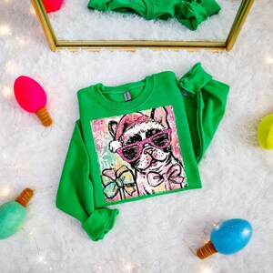 Puede incluir: Una sudadera verde con un gráfico de un bulldog francés con gafas de sol rosas y un gorro de Papá Noel. El perro está rodeado de copos de nieve y lazos. Decoraciones navideñas.