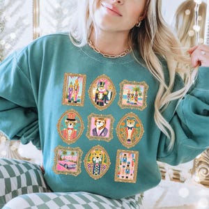 May include: Teal-colored sweatshirt featuring nine framed illustrations of anthropomorphic animals in ornate gold frames. The illustrations include a tiger in a suit, a leopard in a tuxedo, and other whimsical designs. The sweatshirt has a crew neck and long sleeves.
