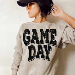Op de afbeelding: Een beige sweatshirt met de tekst "GAME DAY" in grote, vervaagde zwarte letters. De sweatshirt heeft lange mouwen en een ronde hals. De afbeelding toont een persoon die de sweatshirt draagt.