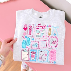 May include: A light gray sweatshirt featuring a colorful graphic design with the word "nurse stuff." The design includes medical-themed illustrations such as a stethoscope, syringe, and bandages, along with hearts and flowers.