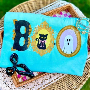 May include: A bright turquoise t-shirt featuring the word BOO in black, with the letter O replaced by two ornate gold frames. One frame contains a black cat with a pink bow, and the other a white ghost. Spiderwebs and spiders decorate the design.
