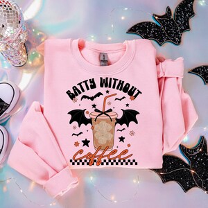 May include: A light pink sweatshirt with the text "Batty Without Coffee" and a graphic of a coffee drink with bat wings. The design includes bats, stars, and flowers. The sweatshirt is folded, with the sleeves visible.