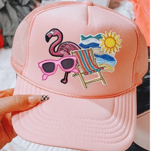 May include: Pink trucker hat with a flamingo, sunglasses, beach chair, sun, and ocean wave patches. The hat has a mesh back and a curved brim. A fun, summery accessory.