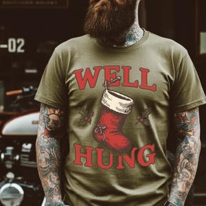 Puede incluir: Camiseta verde oliva con un gráfico de calcetín navideño rojo y las palabras "WELL HUNG" en letras rojas. El calcetín tiene un puño blanco y ramitas de acebo.