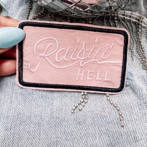 Puede incluir: Un parche rectangular rosa con las palabras "Raisin HELL" bordadas en escritura blanca. El parche tiene un borde negro y está colocado sobre un fondo de mezclilla. Flecos de pedrería cuelgan de la parte inferior.