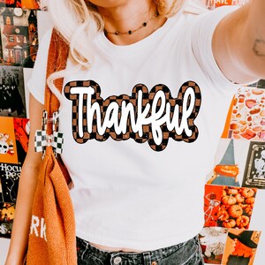 Könnte beinhalten: Weißes T-Shirt mit dem Wort "Thankful" in weißen Buchstaben auf einem braunen und schwarzen karierten Hintergrund.
