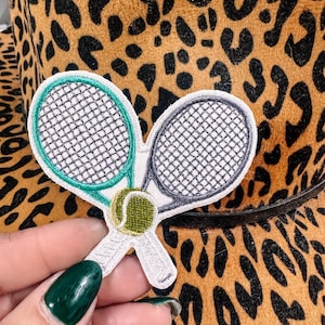 May include: Embroidered patch showing two tennis rackets and a tennis ball. The patch has a white border, with one racket outlined in teal and the other in grey. The tennis ball is green and white. The background is a leopard print.
