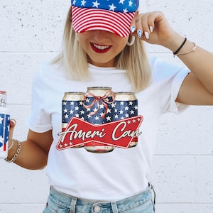 May include: White t-shirt with a graphic of three beer cans. The cans have a blue background with white stars and are tied with a red bow. The text "Ameri Can" is displayed on a red banner. The design has a patriotic theme.