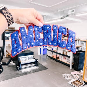May include: A blue chenille patch with white stars spelling the word AMERICA in a bold, block font. The letters are outlined in a thin red border, creating a textured, raised appearance suitable for application onto various fabric surfaces.