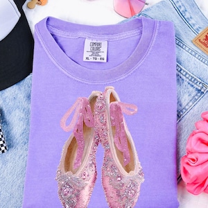 May include: A lilac Comfort Colors t-shirt featuring a pair of pink ballet slippers with beaded embellishments. The slippers have pink ribbons and are adorned with rhinestones. The shirt is size XL-TG-EG.