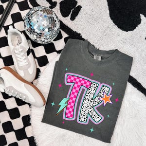 May include: Dark gray t-shirt with a colorful graphic design featuring the letters "TK". The "T" is pink and checkered, while the "K" has a black and white dalmatian print. The shirt is paired with white sneakers and a disco ball.