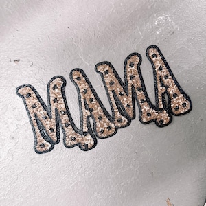 Puede incluir: Un parche decorativo con la palabra MAMA en una fuente estilizada. Las letras están cubiertas de lentejuelas doradas con un estampado de leopardo negro y tienen un borde negro grueso, lo que crea una apariencia audaz y texturizada.