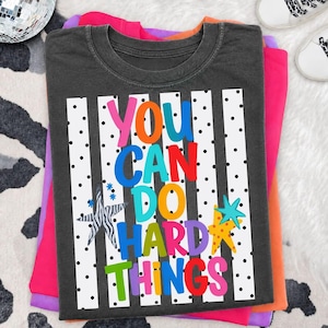 May include: A charcoal gray t-shirt featuring a white rectangular graphic with black vertical stripes and polka dots. The text reads YOU CAN DO HARD THINGS in multicolored letters, accompanied by a zebra-striped star and a solid yellow star.