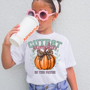 May include: White t-shirt with a graphic of an orange pumpkin with a leopard print bow, the text "Cutest Pumpkin in the Patch", and green leaves. The shirt is worn by a young person wearing pink sunglasses.