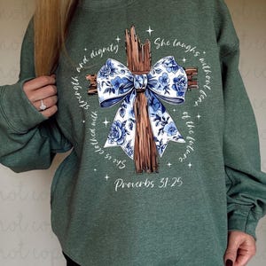 May include: Green sweatshirt with a graphic of a wooden cross adorned with a blue floral bow. The text reads "She is clothed with strength and dignity" and "She laughs without fear of the future" with "Proverbs 31:25" below.