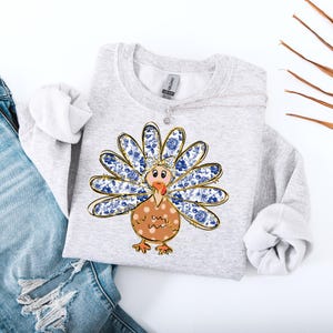 Könnte beinhalten: Ein graues Sweatshirt mit einem blauen und goldenen Truthahn-Design. Der Truthahn hat ein Blumenmuster auf seinen Federn und einen braunen Körper mit weißen Punkten.