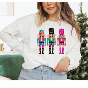Könnte beinhalten: Ein weißer Pullover mit einem festlichen Design mit drei Nussknackern. Die Nussknacker sind mit Glitzer verziert und haben verschiedene farbige Outfits: blau, grün und pink.
