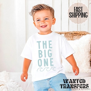 Op de afbeelding: Een wit T-shirt met de tekst "THE BIG ONE" in lichtblauw, met golfafbeeldingen. Het shirt wordt gedragen door een jong kind. De afbeelding bevat ook de tekst "WANTED TRANSFERS DESIGN & APPAREL".