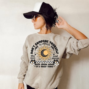 May include: A light beige sweatshirt with a graphic print of a sun and moon with the text "Don't compare yourself to others the sun and moon both shine when it's their time".