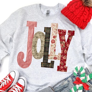 Puede incluir: Una sudadera gris claro con la palabra "JOY" aplicada en varias telas estampadas, incluyendo cuadros rojos, flores y cuadros escoceses. Un gorro de punto rojo y zapatillas rojas también están en la imagen. El tema es festivo y navideño.