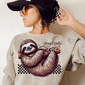 May include: A beige sweatshirt with a graphic of a sloth hanging from a branch. The sloth is brown and has a happy expression. The text "straight chillin'" is printed on the sweatshirt.