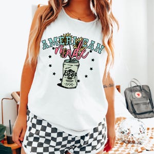 Puede incluir: Camiseta blanca de tirantes con un gráfico de una lata de cerveza con girasoles que crecen de ella. El texto "American Made" está por encima de la lata y el año "1776" está por debajo de la lata.