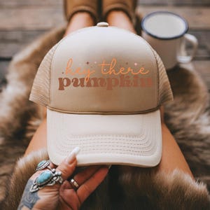 May include: A beige trucker hat with the text "hey there pumpkin" in orange and brown. The hat has a mesh back and a curved brim. A white enamel mug is in the background.