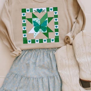 Puede incluir: Una sudadera color beige con un diseño de mariposa verde y blanco, combinada con una falda floral azul claro y botas de vaquero blancas. El diseño de la mariposa presenta una mariposa verde en el centro, rodeada de cuadrados y corazones verdes y blancos.