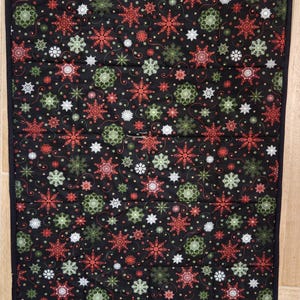 May include: A rectangular quilted fabric runner featuring a black background with a repeating pattern of red, green, and white snowflakes. The runner has a solid black fabric border and a small white rectangular tag in the top left corner.