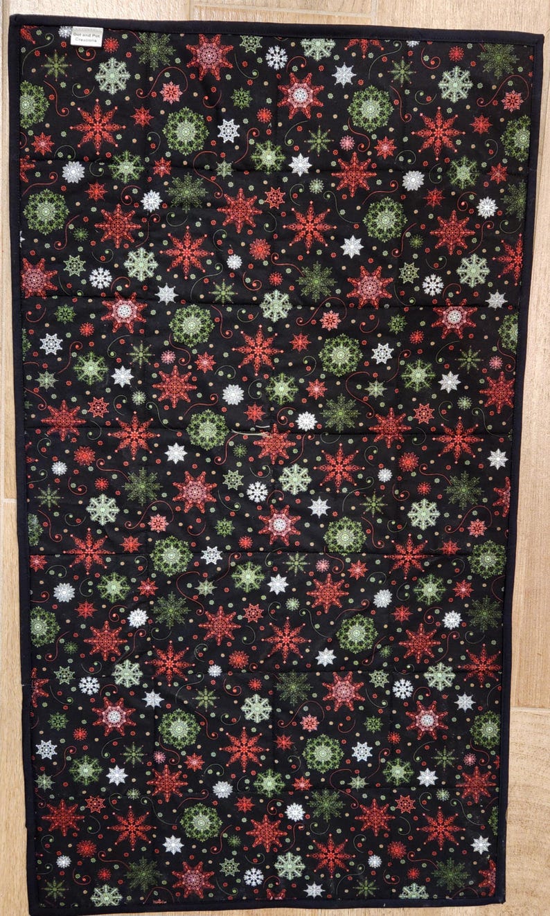 May include: A rectangular quilted fabric runner featuring a black background with a repeating pattern of red, green, and white snowflakes. The runner has a solid black fabric border and a small white rectangular tag in the top left corner.