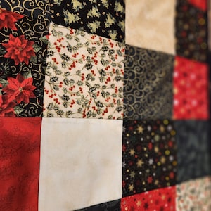 May include: A patchwork quilt featuring a grid of square fabric panels with various holiday-themed prints. The patterns include red poinsettias on black, holly berries and leaves on cream, gold swirls on black, solid red, and gold stars on black.