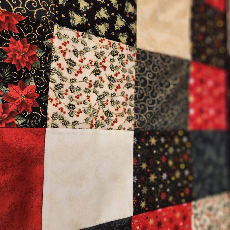 May include: A patchwork quilt featuring a grid of square fabric panels with various holiday-themed prints. The patterns include red poinsettias on black, holly berries and leaves on cream, gold swirls on black, solid red, and gold stars on black.