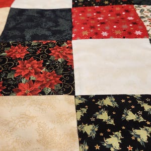 May include: A patchwork quilt featuring a grid of square fabric blocks in various holiday-themed patterns. The design includes red poinsettias on black, white snowflakes on red, cream-coloured holly leaves, and green Christmas trees on black.