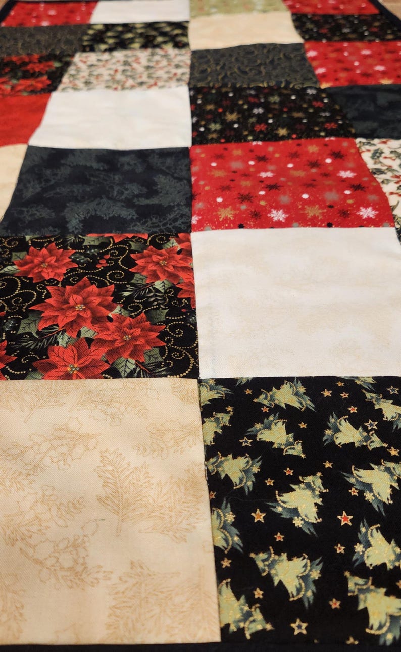May include: A patchwork quilt featuring a grid of square fabric blocks in various holiday-themed patterns. The design includes red poinsettias on black, white snowflakes on red, cream-coloured holly leaves, and green Christmas trees on black.