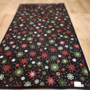 May include: A rectangular fabric table runner featuring a black background with a repeating pattern of red, green, and white snowflakes. The runner has a solid black fabric border around the entire perimeter and a small white rectangular tag.