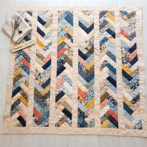 May include: A quilt featuring a herringbone pattern with vertical strips of fabric in shades of blue, yellow, orange, and cream. The quilt has a solid cream border and a wooden tag labeled "Handmade - 52" x 55"" resting on the corner.